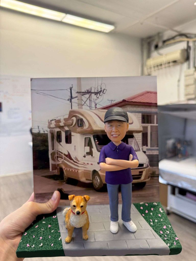 一個訂製的人像公仔退休禮物，公仔穿著紫色上衣和鴨舌帽，雙手抱胸，身旁有一隻可愛的狗，背景是一輛大型露營車和一棟紅色屋頂的房子，地面以灰色人行道磚和綠色草地細膩呈現。