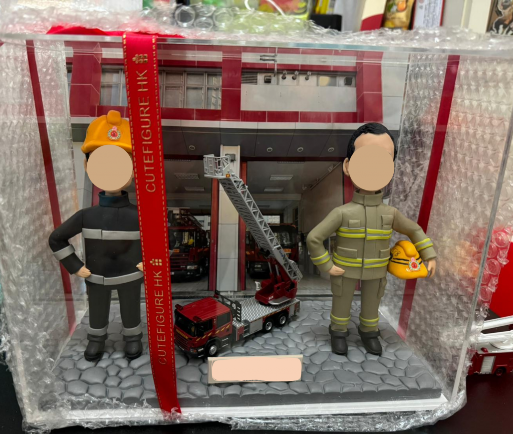 一個精緻的雙人消防員場景模型，放置在帶有CUTEFIGURE HK品牌紅色彩帶的透明展示盒中。模型包含兩位身穿不同款式消防服的公仔，他們的臉部和銘牌均被遮蓋以保護隱私。背景是像真的消防局車房，中央擺放著一輛升降台消防車模型，是一份極具紀念價值的消防員訂製禮物。
