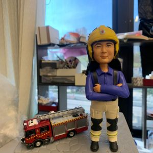 一款為香港消防員訂製的退休人像公仔，公仔為男性消防總隊目，身穿藍色工作服與黃色頭盔，身旁配有一輛紅色消防車模型，由其隊員敬贈。