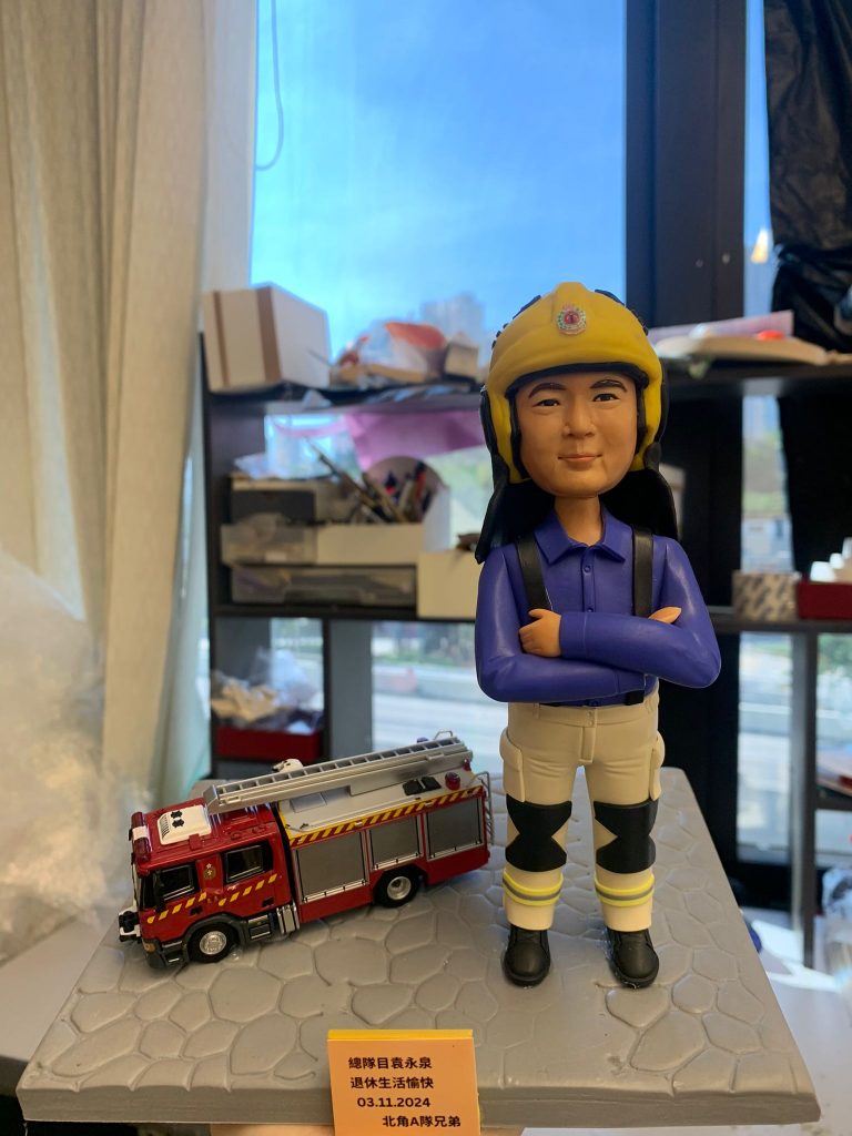 一款為香港消防員訂製的退休人像公仔，公仔為男性消防總隊目，身穿藍色工作服與黃色頭盔，身旁配有一輛紅色消防車模型，由其隊員敬贈。