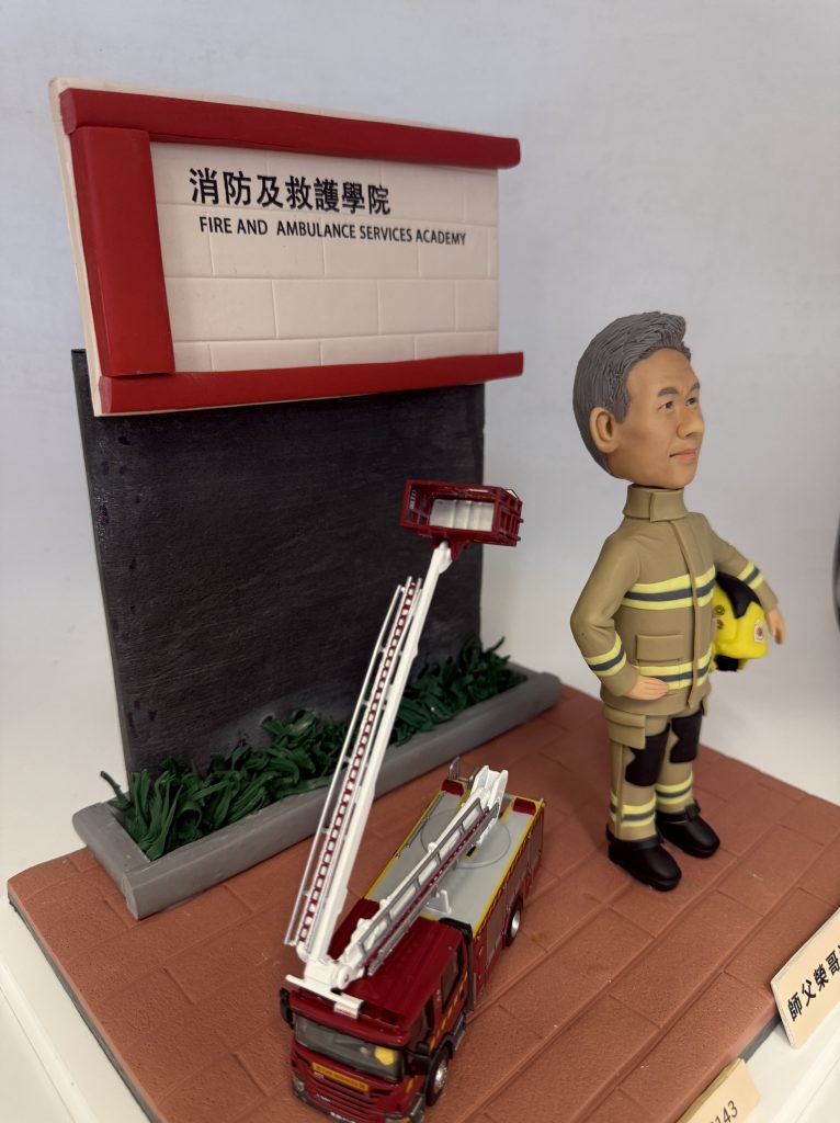 為一位資深消防員訂製的榮休場景模型。模型中，一位有著灰白頭髮的Q版公仔身穿黃金戰衣，站立在「消防及救護學院」的招牌前。他身旁有一輛升降台消防車模型，其雲梯高高升起。這是一份充滿敬意的職業生涯紀念品。