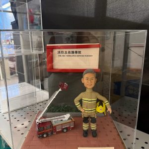 一款消防員榮休禮物，內有一個身穿制服、手持頭盔的人像公仔，背景為消防及救護學院，旁邊有一輛升降台模型車。這份紀念品旨在祝福一位名叫榮哥的師父退休快樂。