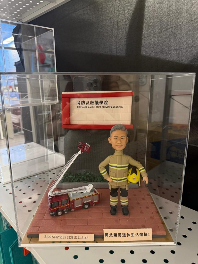 一款消防員榮休禮物，內有一個身穿制服、手持頭盔的人像公仔，背景為消防及救護學院，旁邊有一輛升降台模型車。這份紀念品旨在祝福一位名叫榮哥的師父退休快樂。