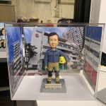 一個消防員的個人化場景模型，放置在透明展示盒中。模型主角身穿藍色襯衫和消防出動褲，手持頭盔。其最大特色是三面環繞的全景相片背景，展示了他所服務的建築群，前方還有一個刻字銘牌。
