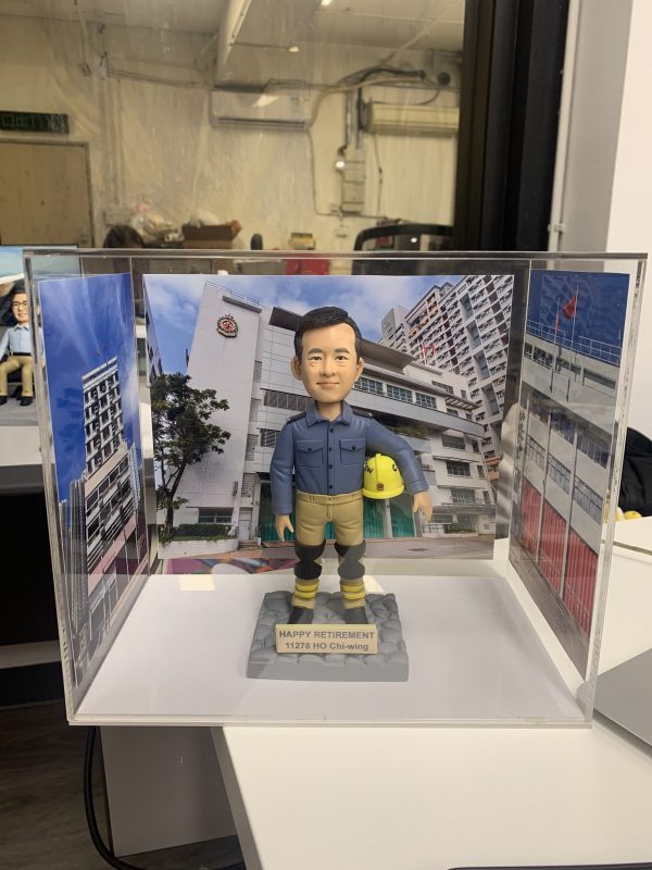 一個消防員的個人化場景模型，放置在透明展示盒中。模型主角身穿藍色襯衫和消防出動褲，手持頭盔。其最大特色是三面環繞的全景相片背景，展示了他所服務的建築群，前方還有一個刻字銘牌。