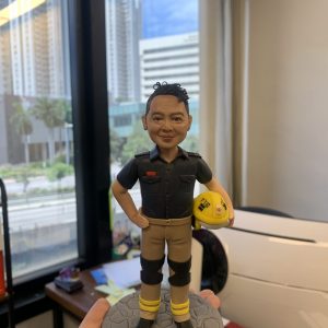一款為香港青衣南消防局C隊的消防人員梁仲斌先生訂製的榮休人像公仔，主角身穿黑色工作制服，手持黃色頭盔，以紀念其光榮退休。