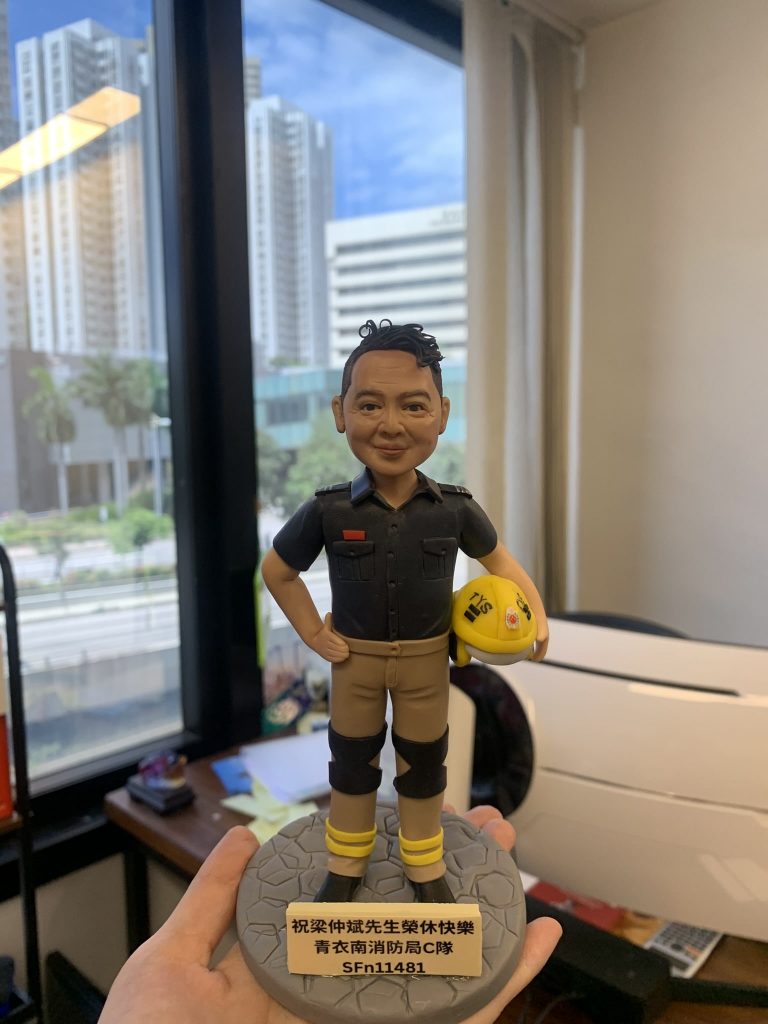 一款為香港青衣南消防局C隊的消防人員梁仲斌先生訂製的榮休人像公仔，主角身穿黑色工作制服，手持黃色頭盔，以紀念其光榮退休。
