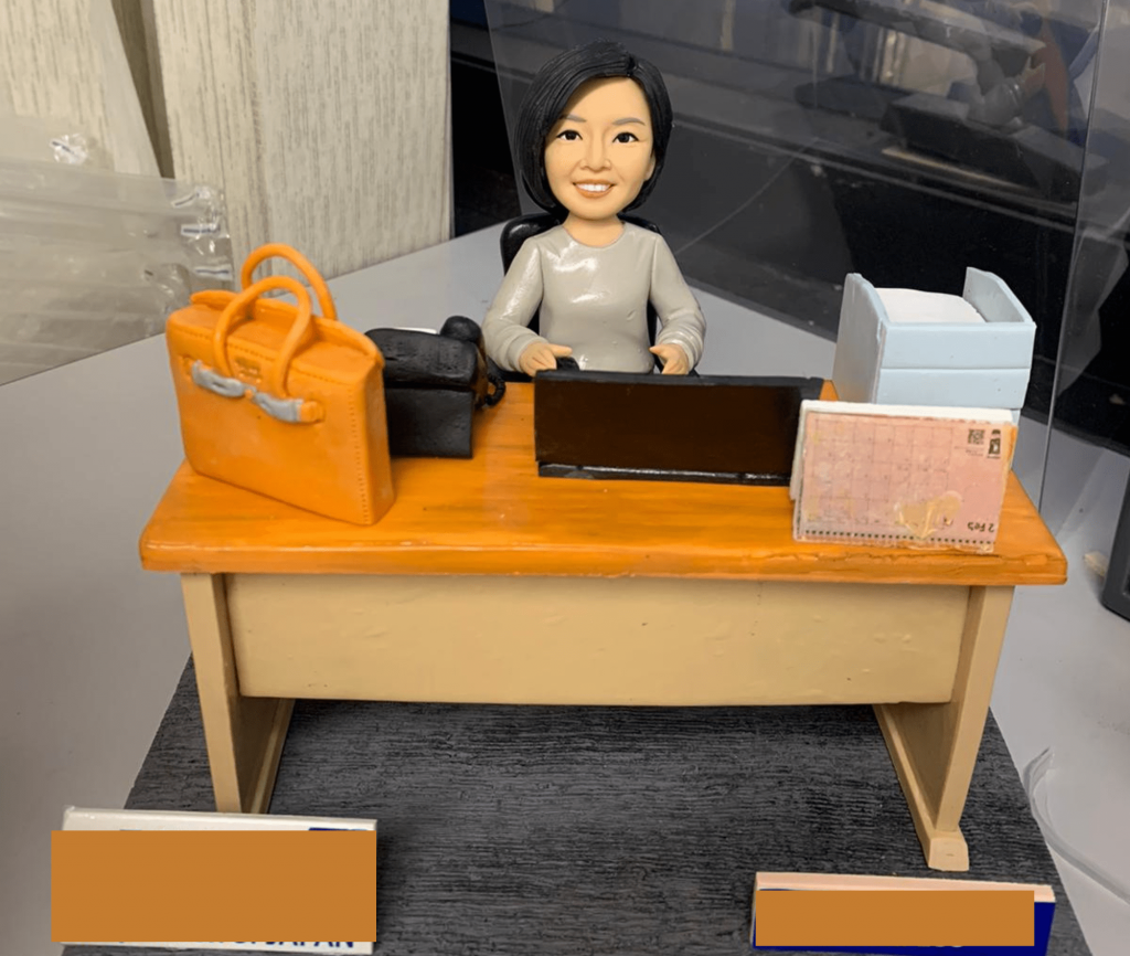 一件為女性上司訂製的精緻人像公仔榮休禮物。留著短髮的公仔主角面帶溫暖微笑，身穿灰色上衣，坐在辦公桌前。桌上最引人注目的是一個亮橙色的名牌手袋，旁邊還有電腦、座檯曆等辦公用品，整個場景生動還原了主角充滿品味的專業日常。
