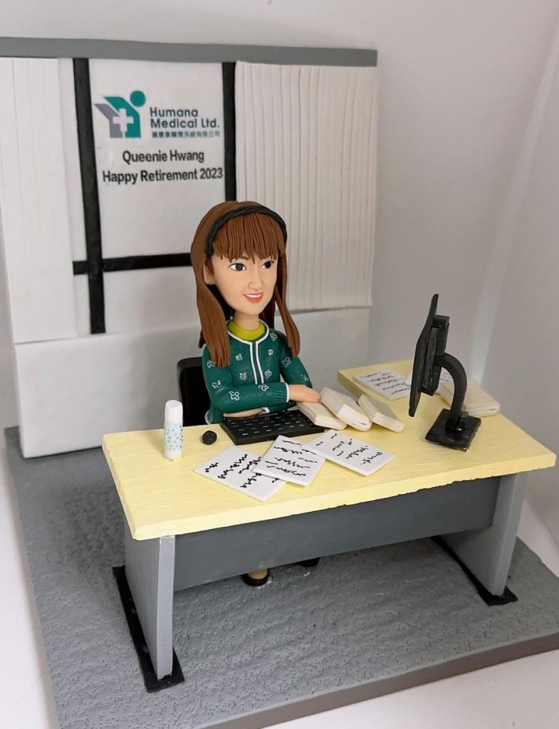 一件為醫療公司員工Queenie Hwang訂製的榮休禮物。一位戴著髮箍的女性人像公仔，面帶微笑地坐在辦公桌前工作。背景牆上有公司「Humana Medical Ltd.」的標誌，並寫著「Queenie Hwang Happy Retirement 2023」，明確表達了這份禮物的祝福。