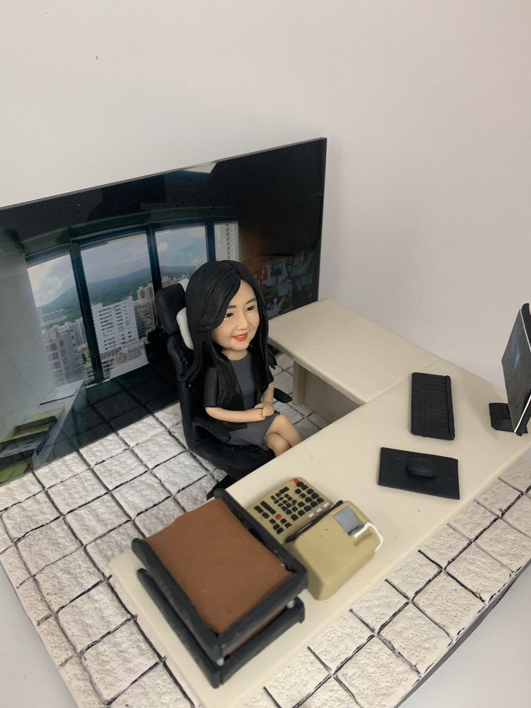 一件極具巧思的女性上司榮休禮物。一位穿著職業裝的女性人像公仔，優雅地坐在她的L形辦公桌前。整個作品最特別的設計在於，背景是一張真實的辦公室窗外風景照片，展現出開闊的城市與山巒景觀，象徵著她所達到的事業高度和她親手打下的江山。