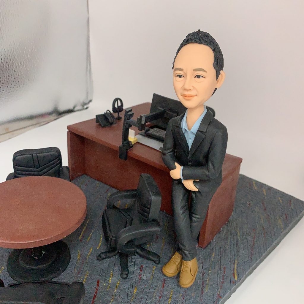 一件送給男性上司的榮休禮物。一位穿著西裝的人像公仔，面帶微笑、雙臂環抱，從容地倚靠在他的行政辦公桌旁。背景是一個完整的高階主管辦公室，除了大班桌和辦公椅，還有一張圓形會議桌和訪客椅，象徵其既是決策者，也是樂於溝通的領導者。