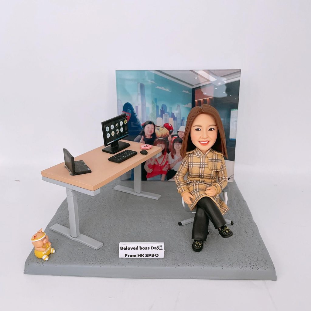 一件極具心思的女性上司榮休禮物。一位穿著黃色格紋風衣的時尚女性人像公仔，面帶微笑地坐在辦公椅上。整個作品最特別的設計在於，背景是一張真實的團隊大合照照片，充滿了溫馨的回憶，象徵著上司與團隊的深厚情誼。底座的牌匾寫著「Beloved boss Da 姐」。