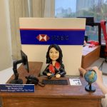 一件為滙豐銀行(HSBC)分行經理 Ms. Maggie Mok 訂製的精緻人像公仔榮休禮物。公仔主角面帶微笑，身穿帶有紅色領巾的專業制服，坐在木紋辦公桌前。背景是一面巨大的HSBC標誌牆，桌上有電腦、地球儀及寫有她姓名職位的銘牌，設計充滿對一位頂級團隊領導的敬意。