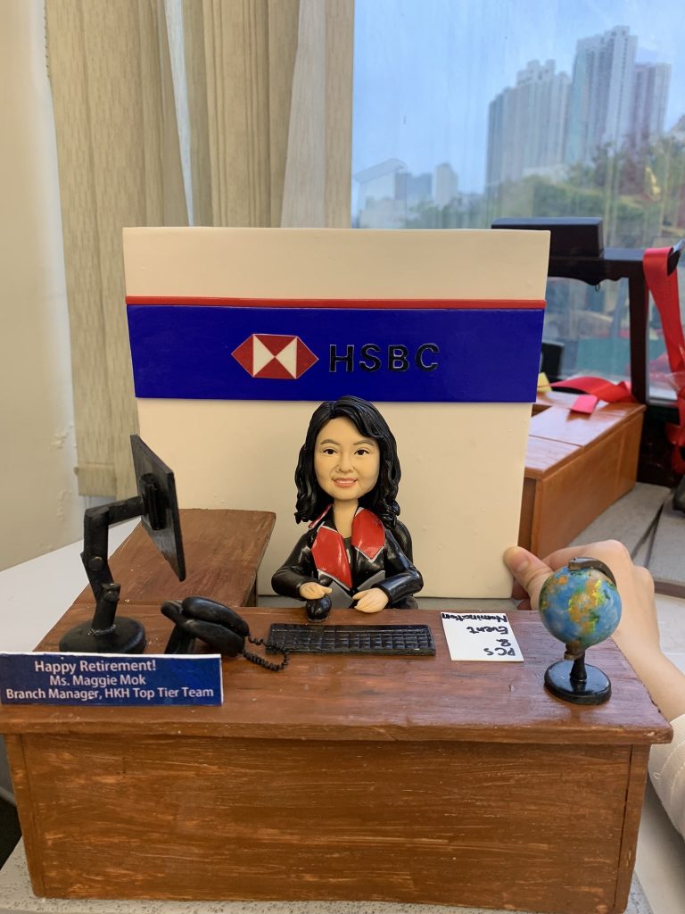 一件為滙豐銀行(HSBC)分行經理 Ms. Maggie Mok 訂製的精緻人像公仔榮休禮物。公仔主角面帶微笑，身穿帶有紅色領巾的專業制服，坐在木紋辦公桌前。背景是一面巨大的HSBC標誌牆，桌上有電腦、地球儀及寫有她姓名職位的銘牌，設計充滿對一位頂級團隊領導的敬意。
