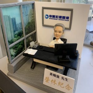 一件為申萬宏源香港的黃熾強先生訂製的精緻人像公仔榮休禮物。銀髮的公仔主角面帶沉穩微笑，安坐於辦公桌前，背景牆上有申萬宏源香港的公司標誌。最引人注目的是他身側有一扇大窗，展現出繁華的都市景觀。作品前方擺放著一塊寫有『黃熾強先生 榮休紀念』的金色字樣銘牌，整體設計充滿故事感與崇高敬意。