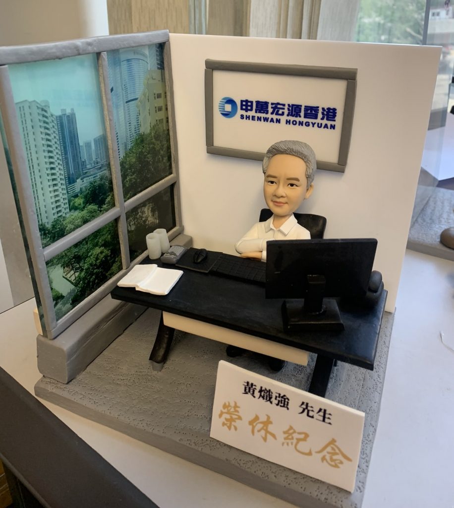 一件為申萬宏源香港的黃熾強先生訂製的精緻人像公仔榮休禮物。銀髮的公仔主角面帶沉穩微笑，安坐於辦公桌前，背景牆上有申萬宏源香港的公司標誌。最引人注目的是他身側有一扇大窗，展現出繁華的都市景觀。作品前方擺放著一塊寫有『黃熾強先生 榮休紀念』的金色字樣銘牌，整體設計充滿故事感與崇高敬意。