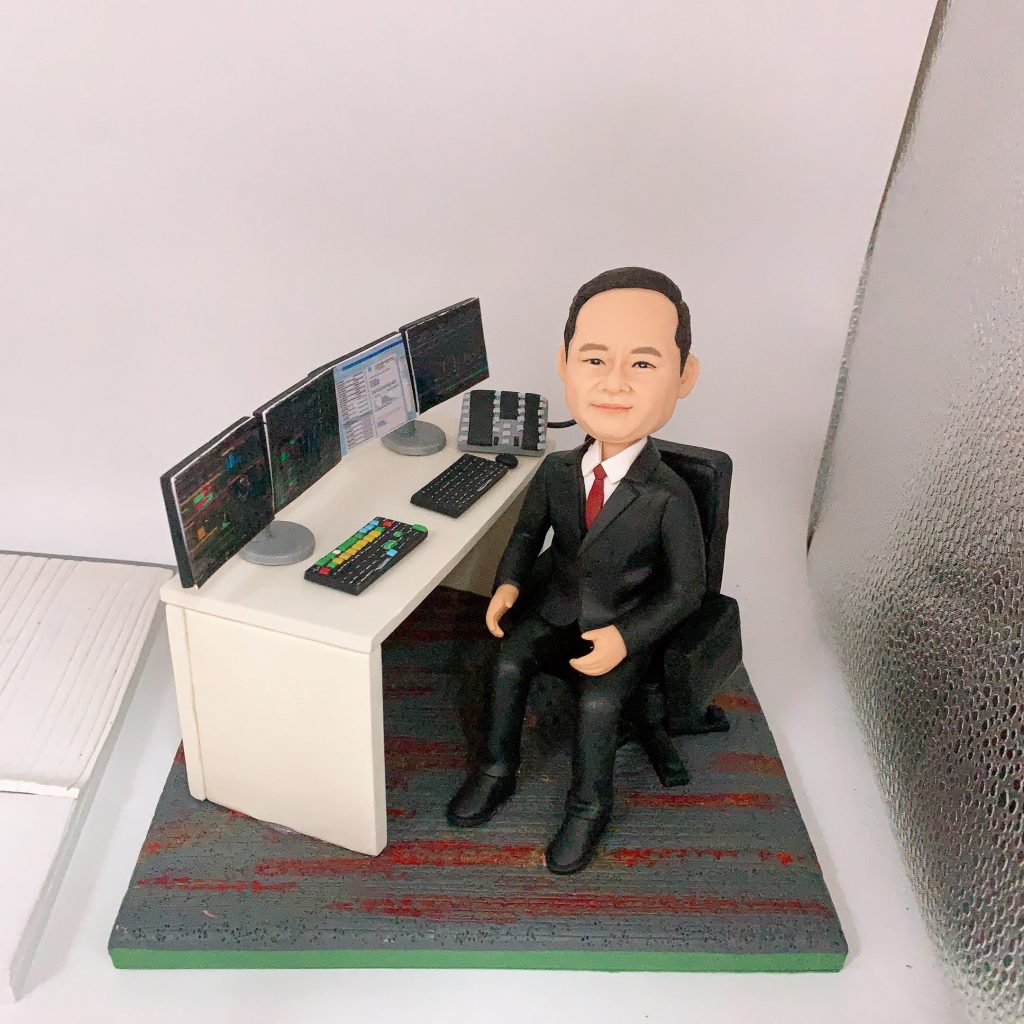 一件為金融界上司訂製的榮休禮物。一位穿著西裝、打紅色領帶的人像公仔，沉穩地坐在辦公桌前。背景極具特色，桌上擺放著一排四個顯示著金融圖表和數據的電腦屏幕，還有一組專業鍵盤，完美還原了交易員或基金經理的工作場景。