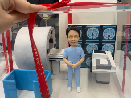一位男醫生的人像公仔，他自信地站在一台精密的磁力共振（MRI）儀器模型旁，背景是腦部掃描的影像，整個場景被包裝在一個繫有紅色絲帶的透明禮物盒中，是一款由康復病人贈送給主診醫生的感謝禮物。