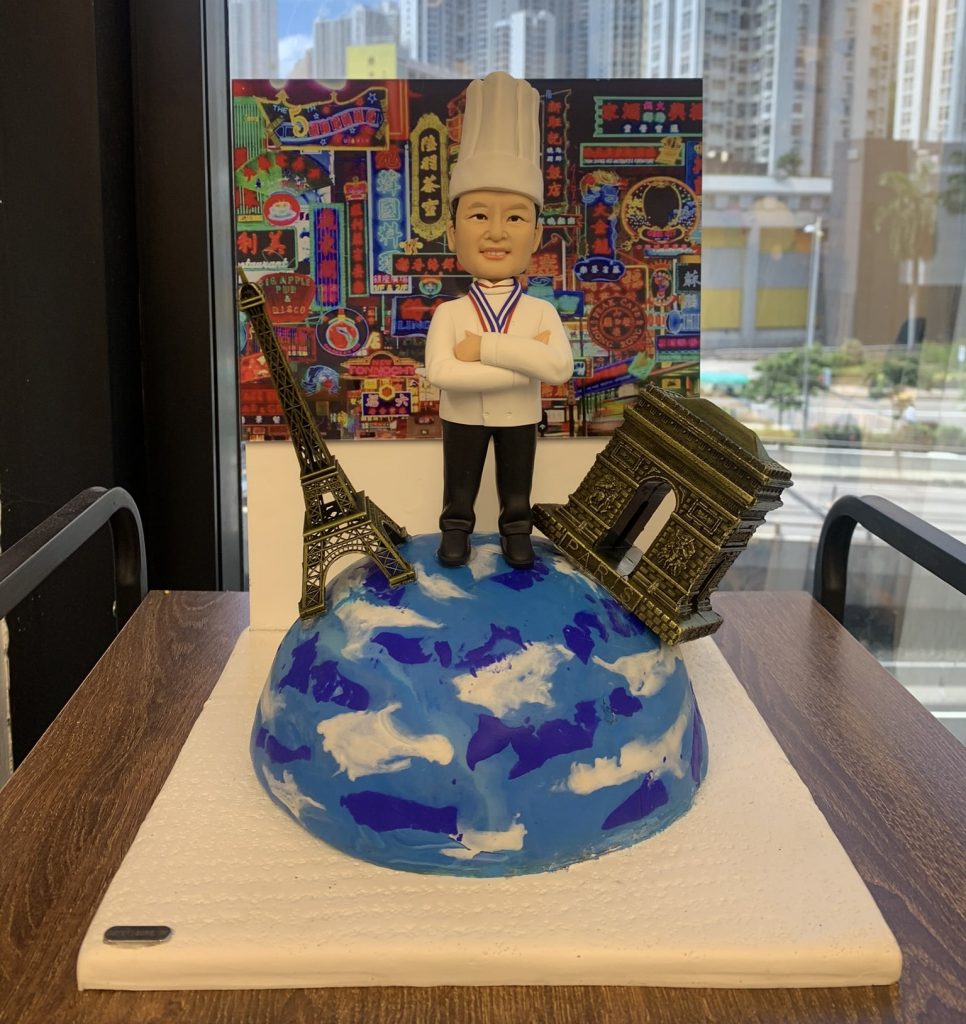 一個為廚師訂製的榮休禮物人像公仔。模型主角是一位穿著廚師服、胸前掛著獎牌的男士，自信地站在地球模型上。地球旁有巴黎鐵塔和凱旋門模型，背景則是一幅充滿香港特色的霓虹燈招牌畫作。整個作品象徵著一位植根香港、在法式烹飪領域取得世界級成就的大廚，是一份極具意義的退休禮物。