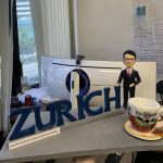 老闆Geoffrey訂製的人像公仔禮物，公仔身穿西裝，自信地站在藍色的「ZURICH」公司名字旁，桌上還有一個咖啡杯，底座刻有團隊的感謝話語，是一份充滿敬意的禮物。