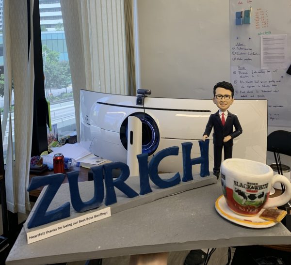 老闆Geoffrey訂製的人像公仔禮物，公仔身穿西裝，自信地站在藍色的「ZURICH」公司名字旁，桌上還有一個咖啡杯，底座刻有團隊的感謝話語，是一份充滿敬意的禮物。