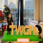 一款為東華三院（TWGHs）成員訂製的人像公仔禮物，描繪了一位身穿黑裙的女士，面帶微笑地站在機構的巨大字母標識旁，底座上還有一個花籃，象徵著她的貢獻與團隊精神。