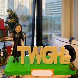 一款為東華三院（TWGHs）成員訂製的人像公仔禮物，描繪了一位身穿黑裙的女士，面帶微笑地站在機構的巨大字母標識旁，底座上還有一個花籃，象徵著她的貢獻與團隊精神。