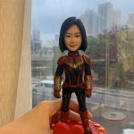 一款手工人像公仔，短黑髮女性面相，穿紅、黑、金配色的超級英雄風格戰衣：胸前有金色星形胸章，肩甲與前臂護甲為紅金色，前臂護甲帶有金色亮片質感；腿部為黑色裝甲線條。公仔由一隻手托起，腳踏紅色心形底座。