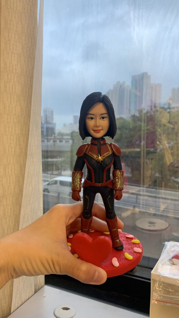 一款手工人像公仔，短黑髮女性面相，穿紅、黑、金配色的超級英雄風格戰衣：胸前有金色星形胸章，肩甲與前臂護甲為紅金色，前臂護甲帶有金色亮片質感；腿部為黑色裝甲線條。公仔由一隻手托起，腳踏紅色心形底座。