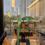 一尊以「督察」職級為主題的警察人像公仔收納於透明壓克力展示盒內，盒蓋繫有綠色緞帶（印字 CUTEFIGURE HK）。公仔短灰黑髮、微笑神情；身穿淺藍色短袖制服襯衫，肩章深色，胸前配有名牌與無線電肩咪走線，腰間見對講機與裝備袋的造型細節；下身為深色長褲與黑色皮鞋。雙臂在胸前交叉，站姿沉穩。腳下為深藍色長方台座，前側豎立白底金字名牌，印有致敬與部門字樣。展示盒內的背板是一個警署內部訓練／會議室場景：多名著制服的人員分排就座、背牆掛有藍底字樣的標語板與投影幕，地面為木紋地板。展示盒外部位於窗邊，窗玻璃上可見雨痕與室內長條燈具倒影；透過窗戶能看到遠處密集住宅高樓與行道樹，畫面左側有手指捏住緞帶的特寫，營造交付禮品的情境。