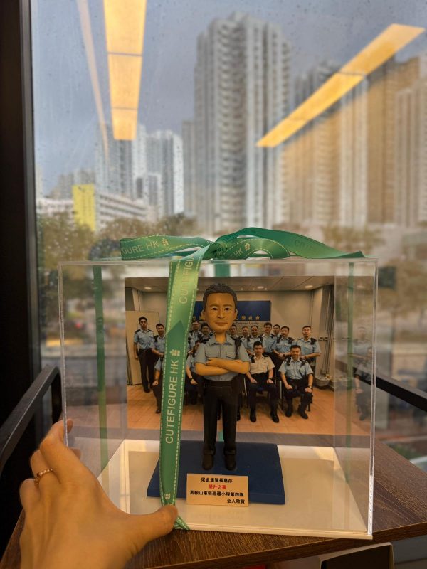 一尊以「督察」職級為主題的警察人像公仔收納於透明壓克力展示盒內，盒蓋繫有綠色緞帶（印字 CUTEFIGURE HK）。公仔短灰黑髮、微笑神情；身穿淺藍色短袖制服襯衫，肩章深色，胸前配有名牌與無線電肩咪走線，腰間見對講機與裝備袋的造型細節；下身為深色長褲與黑色皮鞋。雙臂在胸前交叉，站姿沉穩。腳下為深藍色長方台座，前側豎立白底金字名牌，印有致敬與部門字樣。展示盒內的背板是一個警署內部訓練／會議室場景：多名著制服的人員分排就座、背牆掛有藍底字樣的標語板與投影幕，地面為木紋地板。展示盒外部位於窗邊，窗玻璃上可見雨痕與室內長條燈具倒影；透過窗戶能看到遠處密集住宅高樓與行道樹，畫面左側有手指捏住緞帶的特寫，營造交付禮品的情境。
