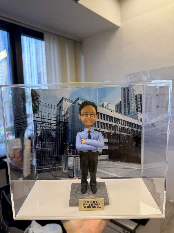 一尊以「警長」職級為主題的警察人像公仔，置於透明壓克力展示盒中，前景由毛絨袖口的手掌托起。公仔短黑髮、戴黑框眼鏡、露齒微笑；身穿淺藍色長袖制服襯衫，肩上有黑色肩章與銀色扣件，領口繫黑色領帶，袖口整齊收邊；下身為深色長褲與黑色皮鞋。公仔雙臂在胸前交叉，姿態沉著自信，站立於灰色六角石紋台座上。台座前方固定金色名牌，刻有祝賀與年份字樣。展示盒後插入的背景板為一座警署建築外觀與鐵欄圍欄的街景，遠處可見高樓大廈與藍天白雲。展示盒外仍能看見室內辦公環境元素，如落地窗邊框、百葉窗簾與牆面，左側窗面反射出長條燈光。整體畫面營造出在職場場景中為「警署警長榮升」而準備的紀念與送禮氛圍。