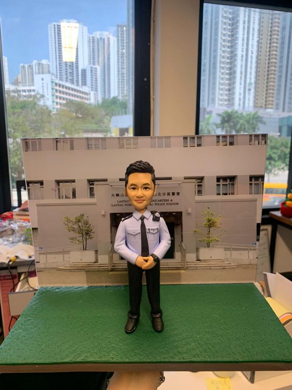 作品以立正合掌的警察人像公仔為主角，透過手作雕刻強調臉部比例、眉眼刻線與唇型弧度，短髮髮束以細筆分層上色呈現光澤；制服部分以淺藍襯衫配黑色領帶與長褲，口袋蓋、鈕扣、肩章和袖口褶位皆以刻紋與陰影處理表現層次，胸前識別章樣式徽片與肩掛對講機組件增添實務感。地台採顆粒化肌理，背板直出警署正門影像，構成「在崗位前敬禮」的情境，既具職務辨識也富儀式感。成品適合作為榮升、退休或嘉許場合的體面贈禮與辦公室展示，亦可客製姓名、職級、頒贈日期與隊章元素，或替換為所屬單位建築背板，使作品更貼近主角故事。