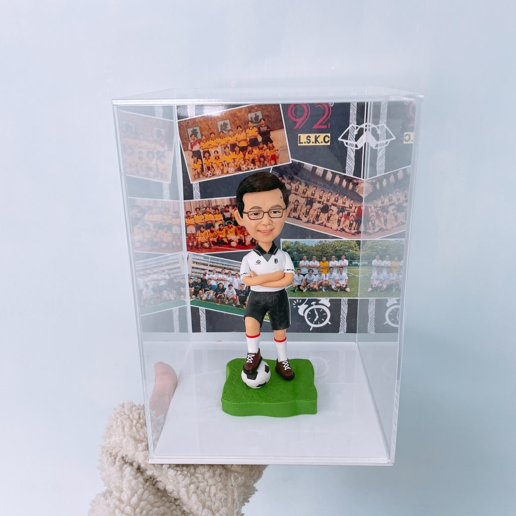 足球教練退休主題人像公仔：戴眼鏡的中年男子公仔身穿白色翻領教練上衣、黑色短褲與棕色球鞋，雙手抱胸自信站立；右腳踩在黑白足球上，腳下為綠色草地造型底座。整體置於透明展示盒內，背景板拼貼多張球隊合照與「92 L.S.K.C」等字樣，盒外可見毛絨外套手掌托住展示盒，象徵向資深足球教練致敬的退休禮物人像公仔。