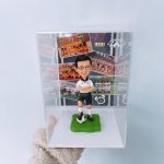 足球運動人像公仔，身穿白色球衣與黑色短褲，雙手交叉自信站立在足球上，擺放於透明展示盒內，背景為球隊合照。適合作為運動紀念、足球愛好者專屬收藏與禮物。