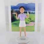 一款客製化人像公仔，呈現一位身穿紫色高爾夫球上衣與白色裙子的女性，肩上揹著高爾夫球桿，臉上帶著笑容。公仔底座寫有「Happy Retirement 2021 from HN Landmark」，背後襯托青山與翠綠草地的高爾夫球場場景，象徵運動、高爾夫球與專屬禮物。