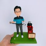 一款客製化人像公仔，呈現一位身穿藍黑拼色運動上衣與灰色褲子的男性，手持高爾夫球桿，腳邊有一顆高爾夫球，旁邊放置紅黑配色的球袋與祝賀牌。公仔以綠色草地為底座，展現高爾夫球運動氛圍，適合作為獨特的運動紀念禮物或退休禮品。