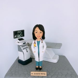 女性醫生人像公仔，身穿白袍與藍綠色醫護制服，脖子掛著聽診器，站在診症室中。背景配置超聲波（Ultrasound）檢查儀與螢幕，上方顯示胎兒影像，旁邊有檢查床，完整呈現婦產科或內科檢查場景。此專屬客製公仔適合作為醫生禮物、醫護人員升職賀禮、退休紀念或醫療團隊感謝禮物。