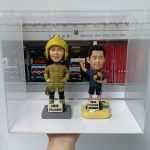 一款為消防員訂製的退休人像公仔禮物，包含兩個公仔，一個穿消防制服，另一個穿休閒服，背景是上環消防局照片，裝在透明保護盒中。