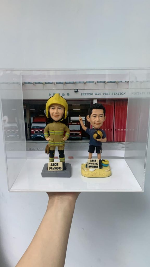一款為消防員訂製的退休人像公仔禮物，包含兩個公仔，一個穿消防制服，另一個穿休閒服，背景是上環消防局照片，裝在透明保護盒中。