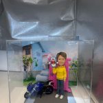 一個人像公仔的展示，公仔穿著黃色外套，手持一個叮噹（多啦夢）公仔，站在一輛藍色摩托車旁。背景是一座現代化的房子與綠意盎然的花園，整個展示在透明的展示盒中。