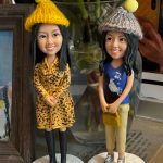 兩個手工製作的人像公仔，細緻還原女生的五官與穿搭，其中一位穿著黃色豹紋洋裝搭配黑色長靴，另一位穿著藍色上衣與淺色長褲，手持小手袋。兩個公仔都戴著可愛的毛線帽，非常適合作為聖誕節禮物或特別紀念品。