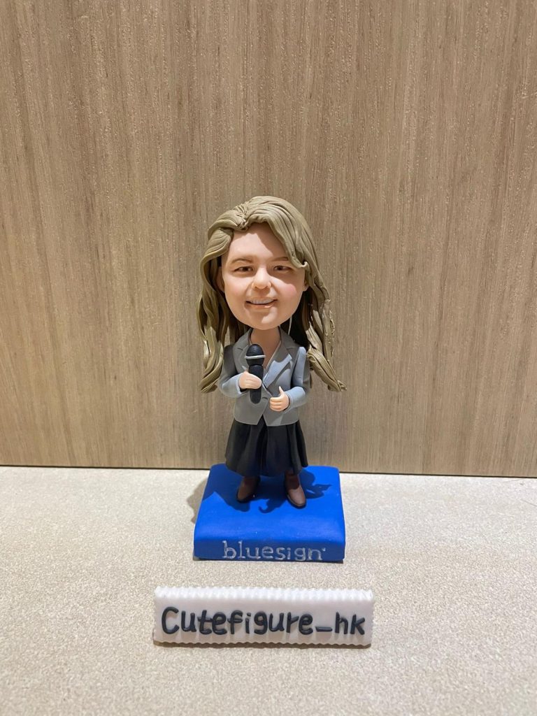 一位金髮女士的專業演講者人像公仔，她身穿灰色西裝外套，手持咪高峰，並站立在一個印有 "bluesign" 公司標誌的藍色地台上。