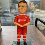 一個穿著紅色利物浦足球隊球衣的人像公仔，面帶微笑，站在綠色草地底座上。公仔穿著利物浦球衣，帶有俱樂部的標誌、Nike標誌，並佩戴紅色的球襪和黑色球鞋。背景為模糊的城市景觀，突出顯示球員的專屬紀念樣式。