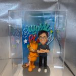 一個人像公仔和加菲貓的塑像展示在透明展示盒中。公仔穿著藍色T恤和黑色褲子，手臂交叉，旁邊的加菲貓站在灰色底座上，也同樣交叉手臂。背景有色彩繽紛的“Go Work”字樣，並搭配星星和閃電的裝飾，營造活潑氛圍。