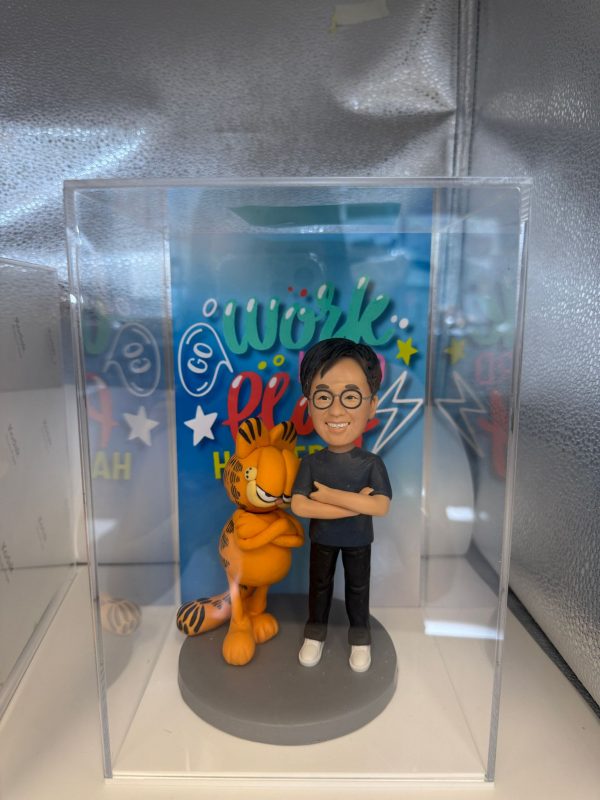 一個人像公仔和加菲貓的塑像展示在透明展示盒中。公仔穿著藍色T恤和黑色褲子，手臂交叉，旁邊的加菲貓站在灰色底座上，也同樣交叉手臂。背景有色彩繽紛的“Go Work”字樣，並搭配星星和閃電的裝飾，營造活潑氛圍。