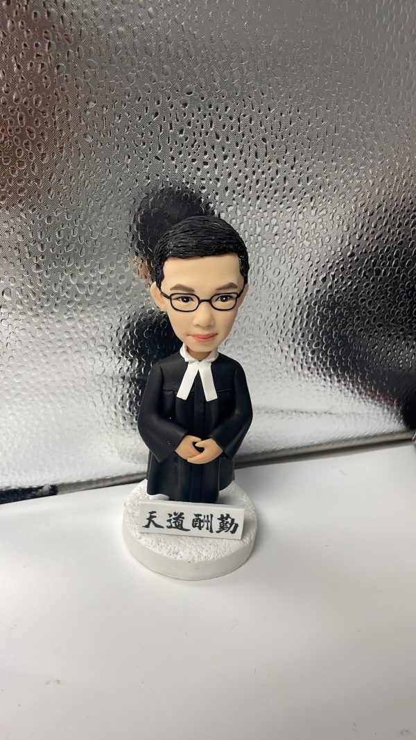 一款男性律師人像公仔，身穿律師袍，站立在刻有「天道酬勤」銘牌的底座上，是送給律師的完美入職禮物。