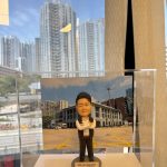 一尊以「副警司」職級為主題的警察人像公仔，收納於透明壓克力展示盒內，由下方手掌托起拍攝。公仔黑色短髮向後梳整，眉眼立體，微帶笑意；穿白色立領襯衫，領口未打領帶，外搭深色無袖戰術背心，肩位與前胸處見立體口袋與按扣造型，兩側配有對講機與肩咪等裝備造型。下身為深色長褲與黑色鞋。雙臂在胸前交叉，呈沉著自信的站姿。公仔站於灰色圓形台座，前側米金色名牌印有英文職銜與部門代碼。展示盒內的背景板是一座具柱廊與拱門語彙的警署建築，前方為畫滿黃色交叉線的停車廣場；遠處可見中高樓建築。展示盒外透過大面窗戶可見密集住宅高樓與藍天白雲，窗面反射出室內燈帶與窗簾邊緣，營造辦公室窗景與城市天際線交錯的視覺層次。