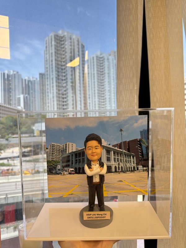 一尊以「副警司」職級為主題的警察人像公仔，收納於透明壓克力展示盒內，由下方手掌托起拍攝。公仔黑色短髮向後梳整，眉眼立體，微帶笑意；穿白色立領襯衫，領口未打領帶，外搭深色無袖戰術背心，肩位與前胸處見立體口袋與按扣造型，兩側配有對講機與肩咪等裝備造型。下身為深色長褲與黑色鞋。雙臂在胸前交叉，呈沉著自信的站姿。公仔站於灰色圓形台座，前側米金色名牌印有英文職銜與部門代碼。展示盒內的背景板是一座具柱廊與拱門語彙的警署建築，前方為畫滿黃色交叉線的停車廣場；遠處可見中高樓建築。展示盒外透過大面窗戶可見密集住宅高樓與藍天白雲，窗面反射出室內燈帶與窗簾邊緣，營造辦公室窗景與城市天際線交錯的視覺層次。