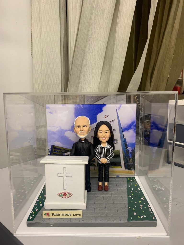 一款客製化牧師人像公仔，呈現一位牧師與女同工站在學校講台後，背景是校園建築，講台有十字架與校徽，是紀念牧師在校服事的榮休感謝禮物。